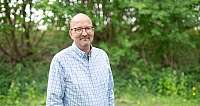 Ein lächelnder Mann mit Brille und kariertem Hemd steht vor grüner Natur