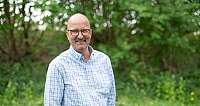 Ein lächelnder Mann mit Brille und kariertem Hemd steht vor grüner Natur