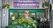 Frau hält Tablett mit jungen Pflanzen vor Gartencenter mit blumenhängenden Töpfen