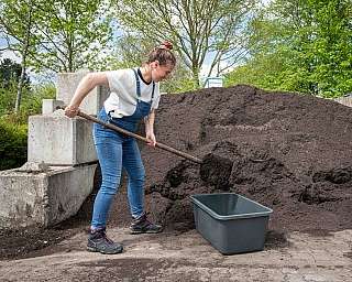 Frau in Overalls füllt Schubkarre mit Erde aus großem Erdbehälter im Freien bei Tageslicht mit Bäumen im Hintergrund, Haustier nicht sichtbar oder vorhanden, Frau trägt Arbeitsschuhe und T-Shirt in heller Umgebung mit blauem Himmel und grünen Blättern am 